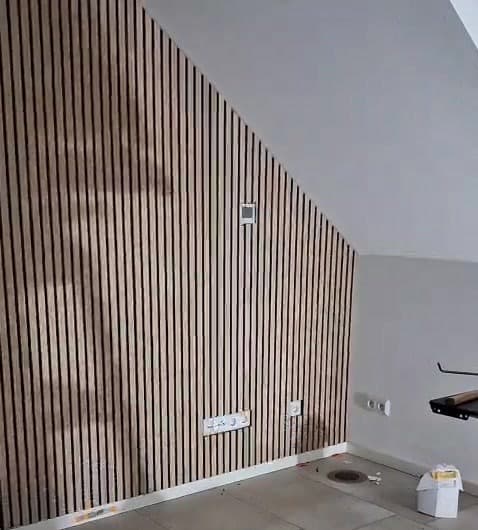 Wand mit senkrechten Holzlamellen unter einer Dachschräge, Steckdosen und ein kleines Wandgerät in die Verkleidung eingepasst